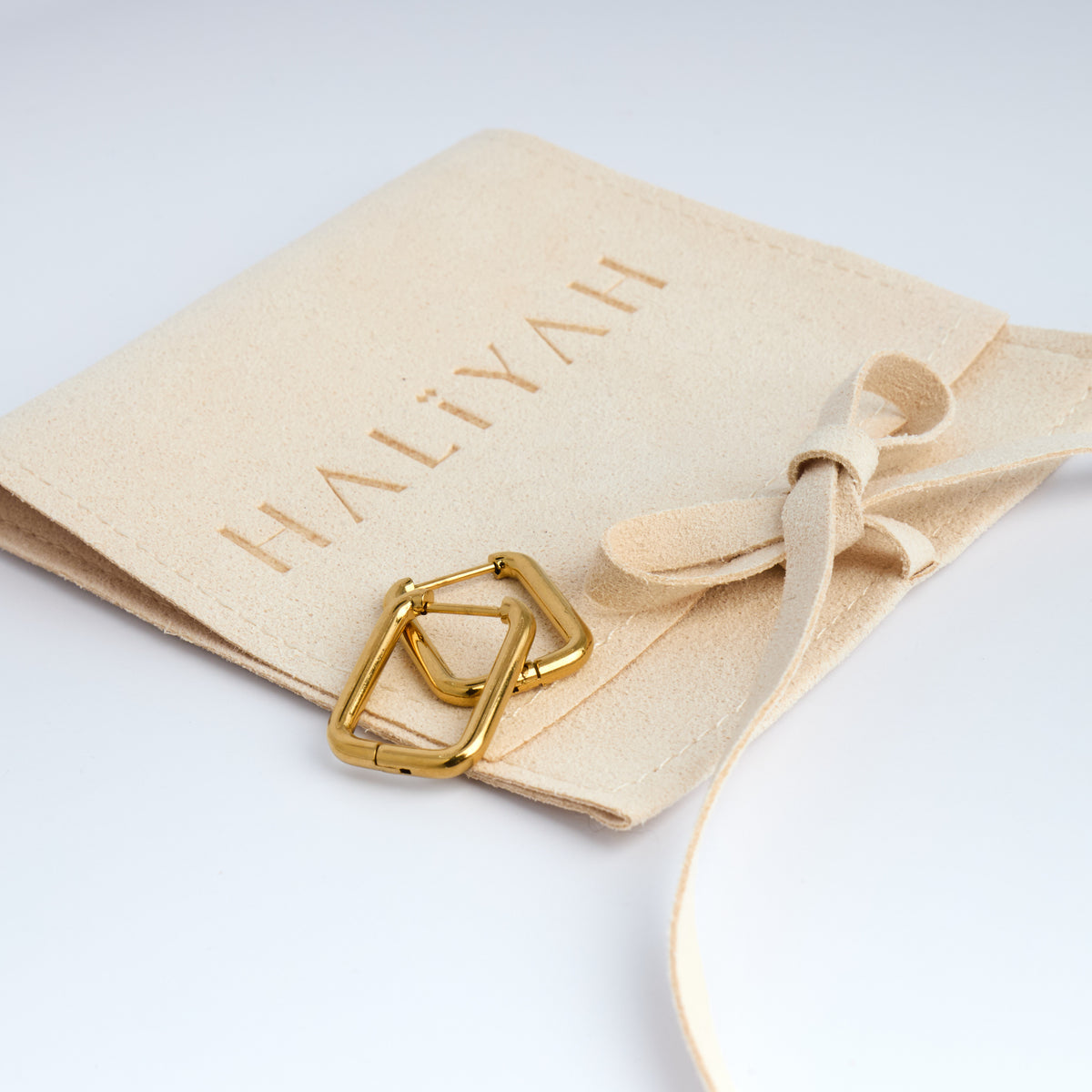 Gold Square Design hoops on a beige drawstring bag with 'HALIYAH' branding.