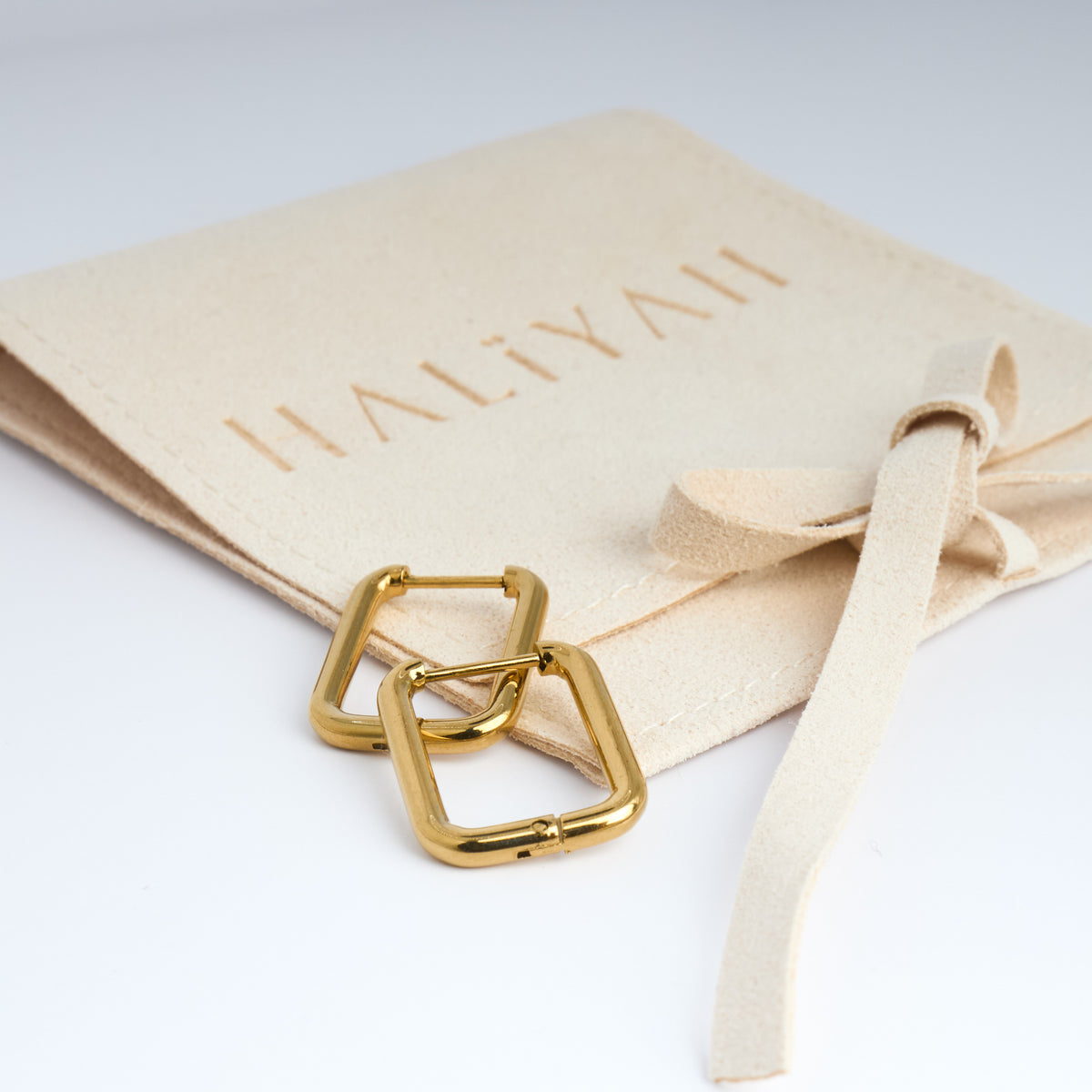 Gold square ring earrings on a beige pouch with 'HALIYAH' branding.